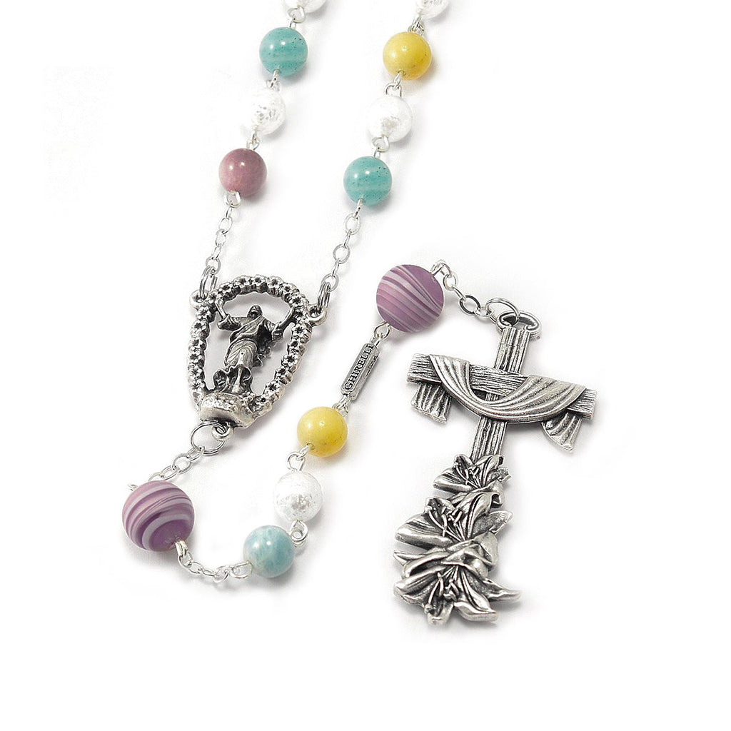 A colorful rosary with white, blue, and yellow beads, featuring a Murano glass crucifix and a lily centerpiece, finished with an antique silver signature bar.